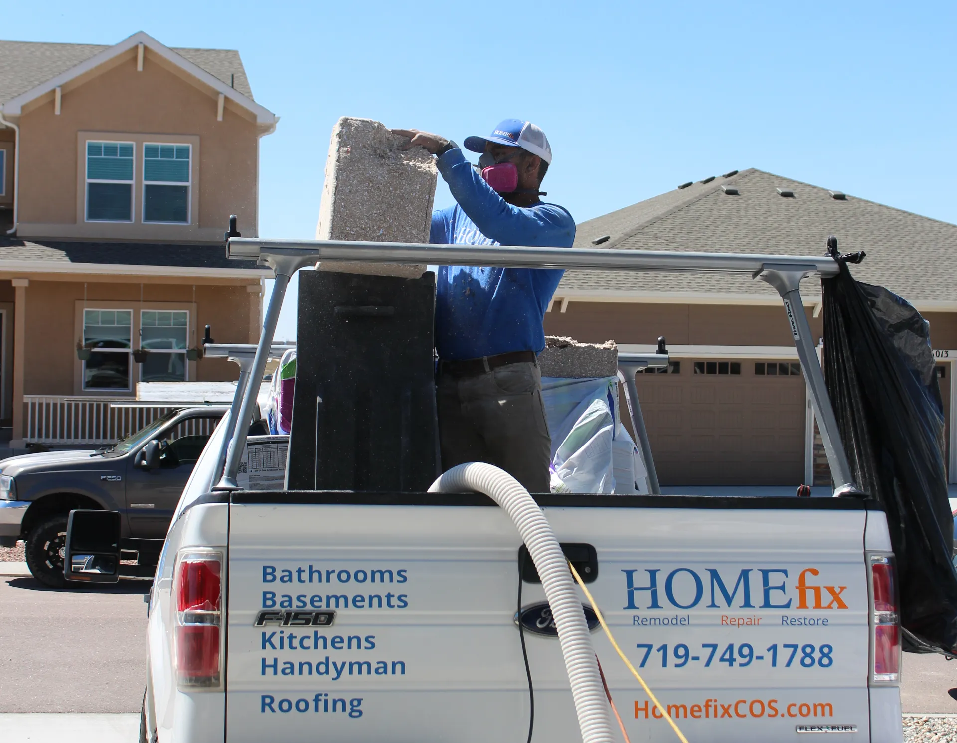 Worker wearing a mask and cap loading insulation material into a white Homefix service truck in a residential neighborhood.