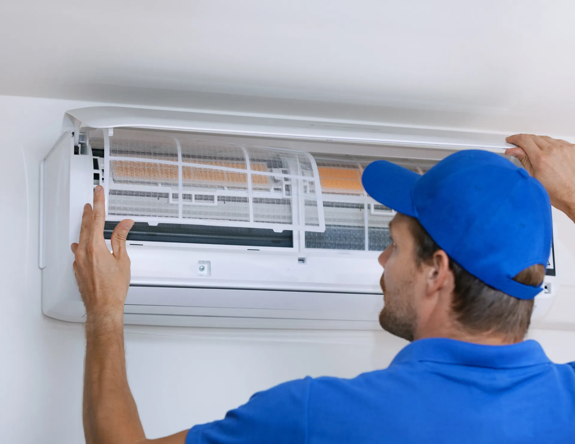 Technician in blue cap and shirt inspecting or servicing a wall-mounted air conditioning unit.