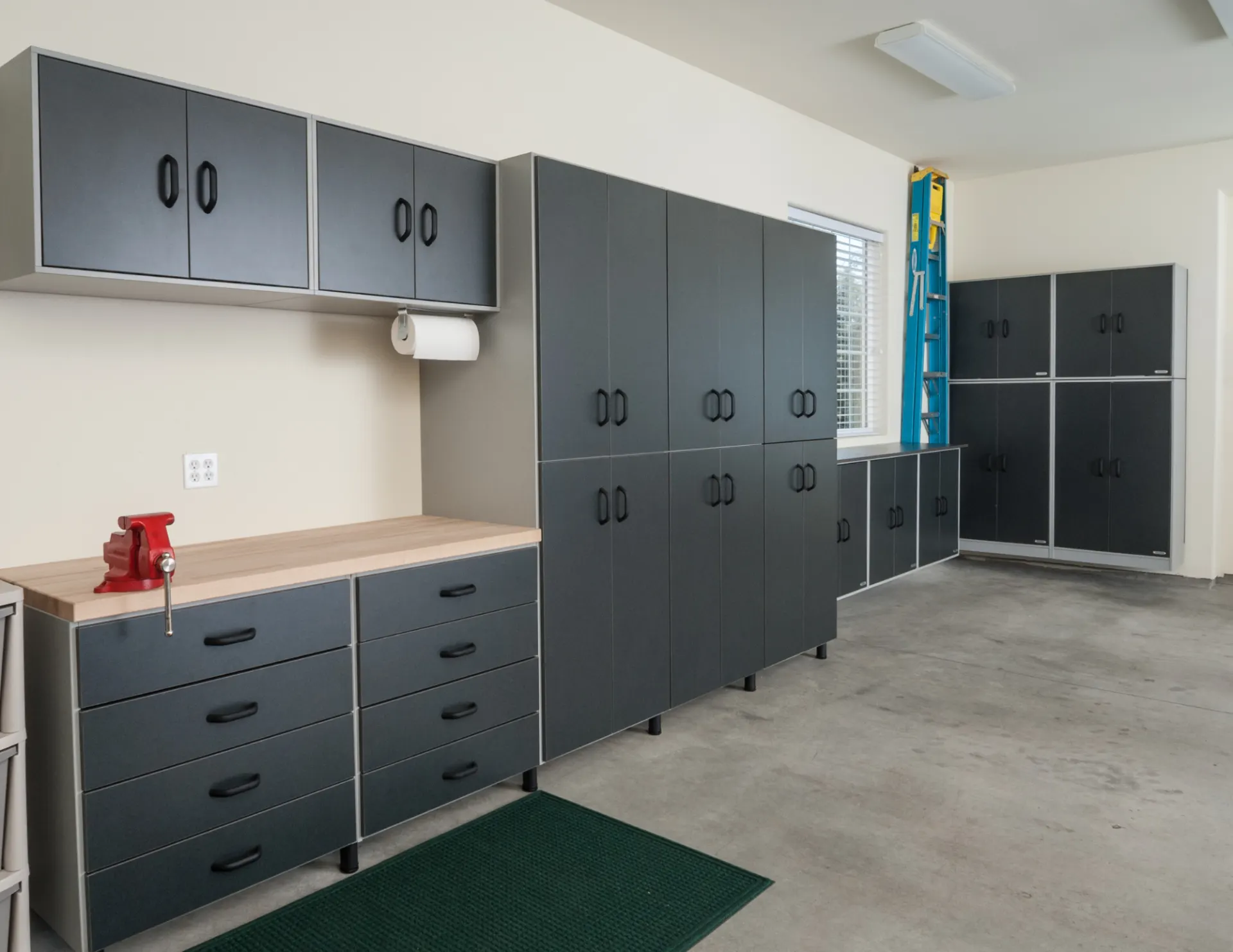Organized garage with black storage cabinets, drawers, a wooden workbench topped with a red vise, and a blue ladder near a window.