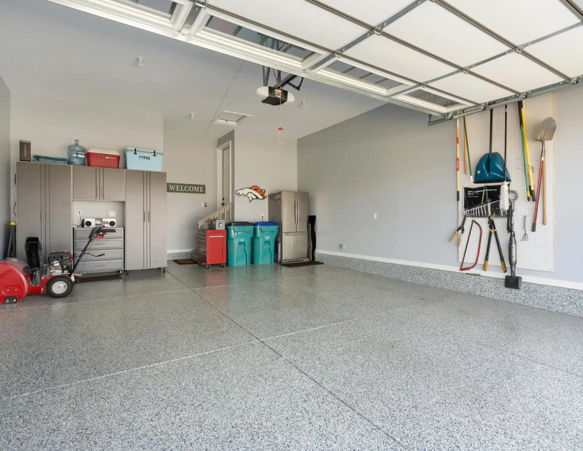 Clean organized garage with polished floor, gray cabinets, red tool chest, recycling bins, and garden tools hanging on a pegboard.