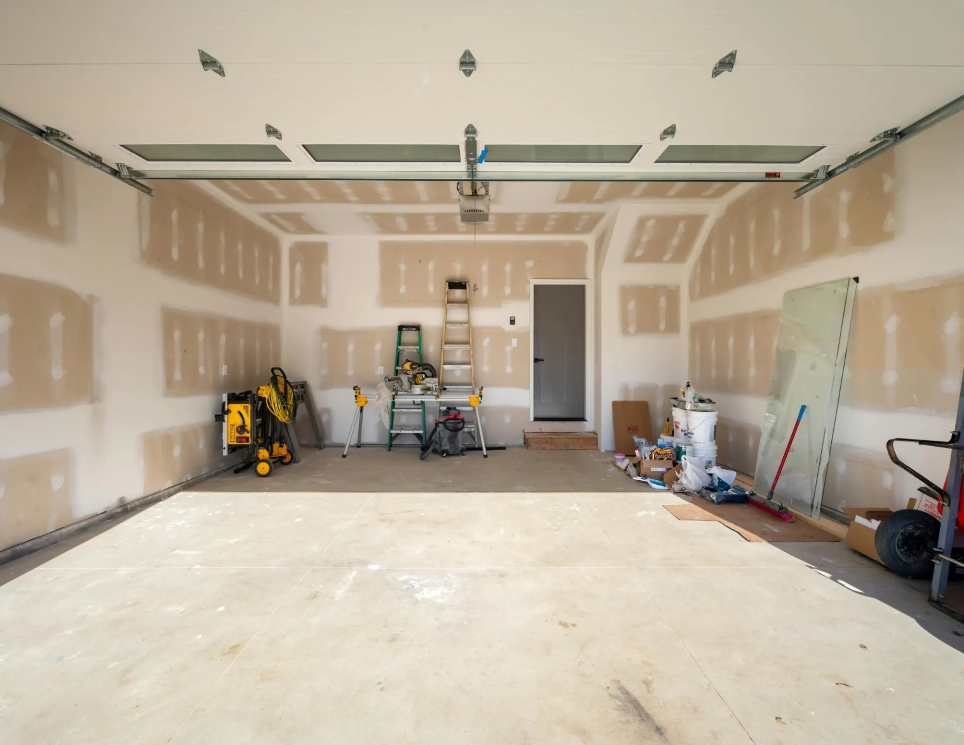 Unfinished garage interior with drywall, construction tools, ladders, and building materials scattered around.