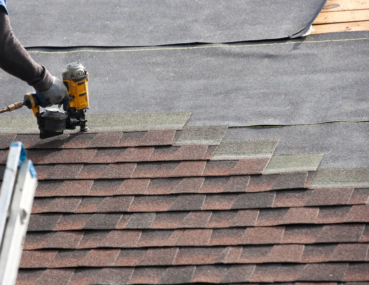 Worker using a nail gun to install asphalt roof shingles on a roof with black underlayment visible.