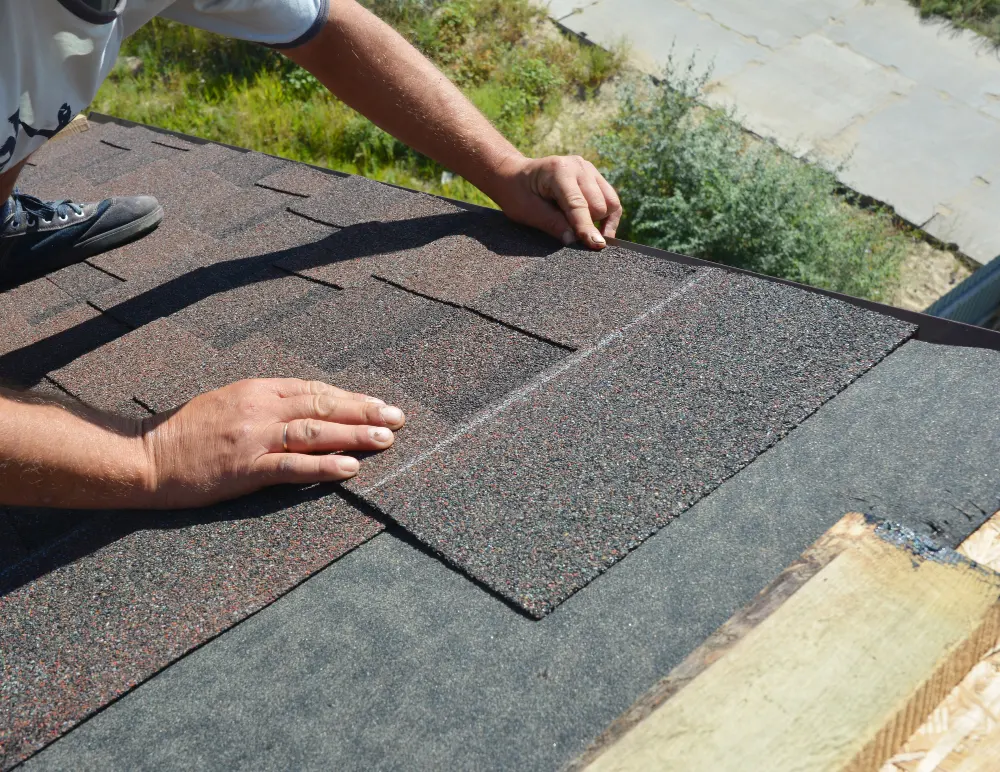 Person installing brown asphalt shingles on a roof under bright daylight.