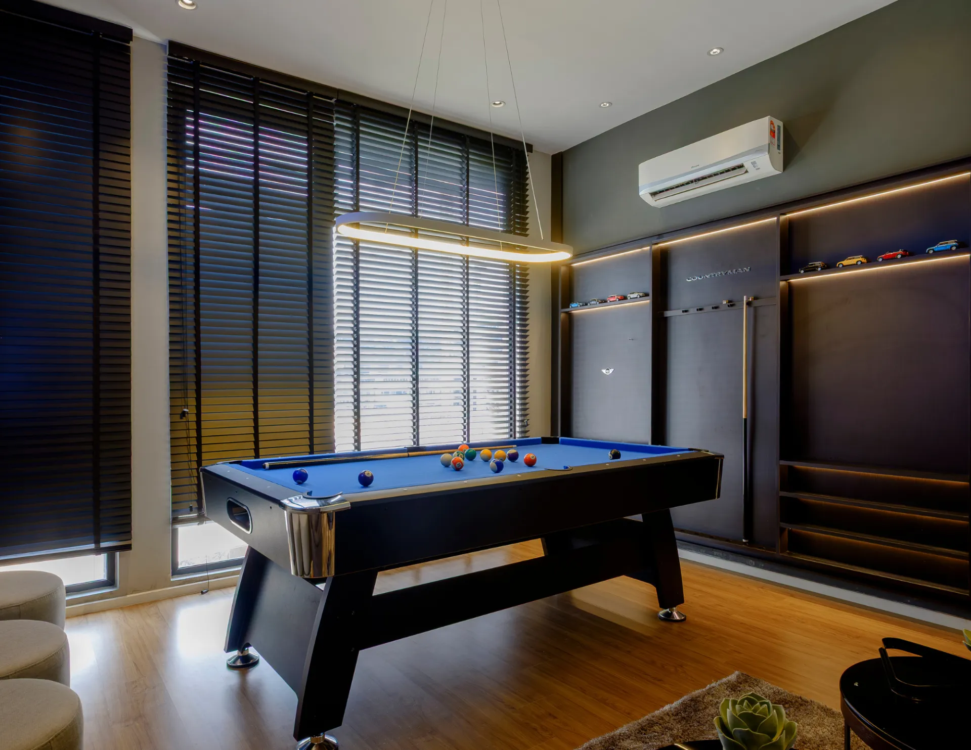 Modern game room with a blue felt pool table, hanging light fixture, and shelves displaying miniature cars on a gray wall with wood flooring.