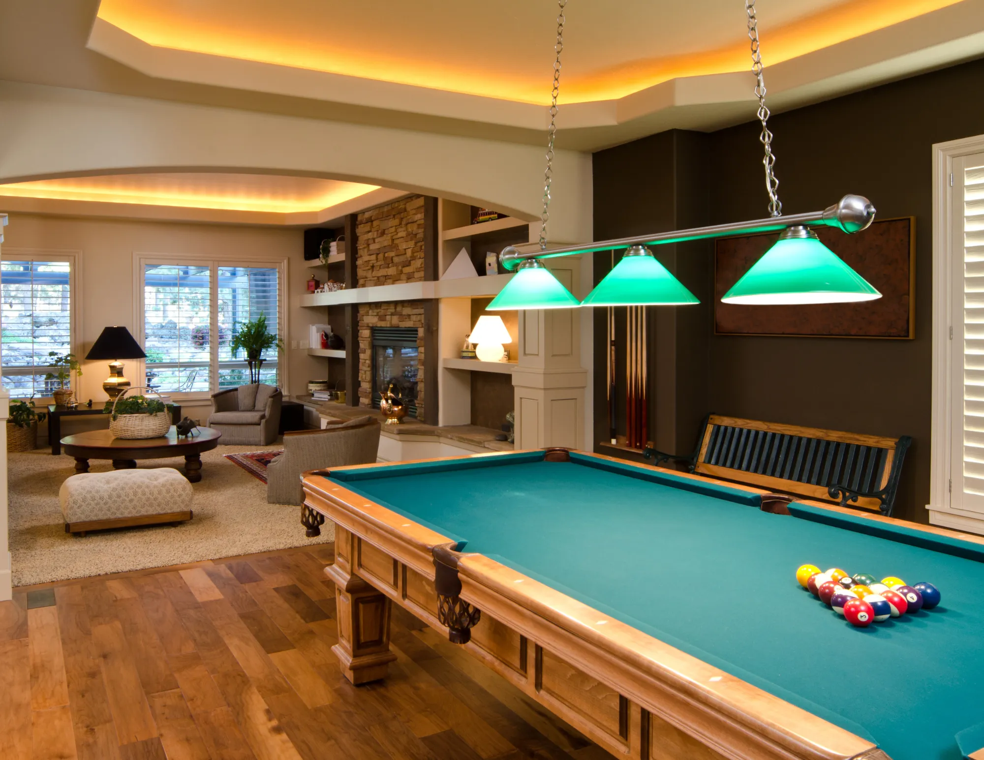 Cozy game room with a pool table under green hanging lights, wooden floor, and a sitting area with armchairs and a fireplace.