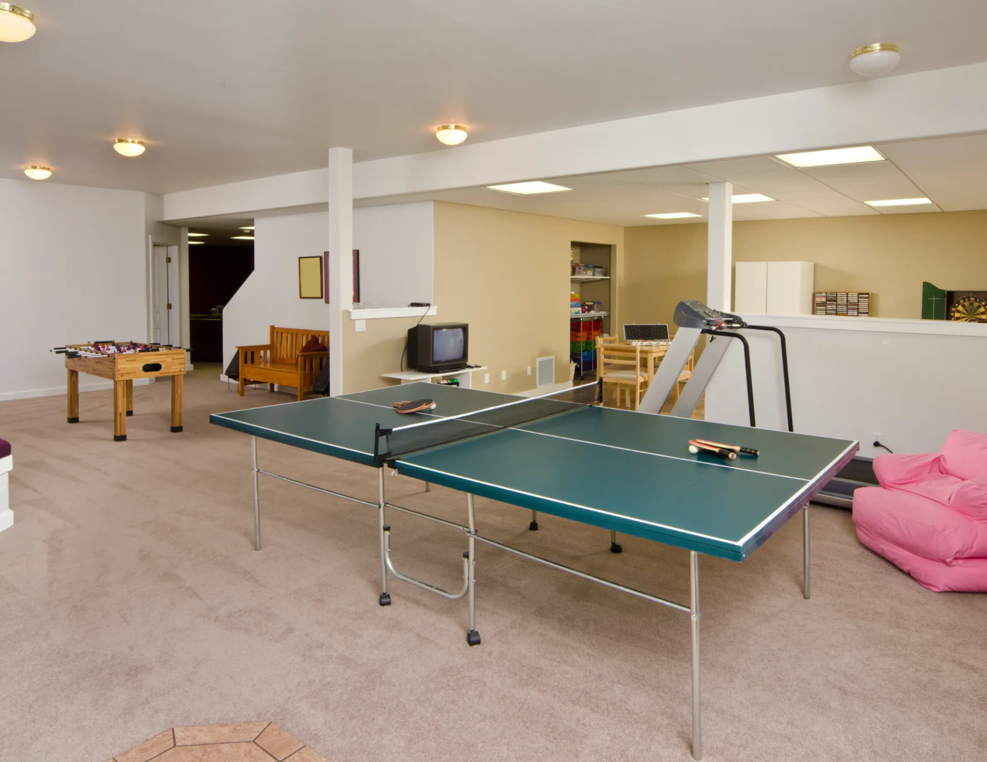 Spacious game room featuring a ping pong table, foosball table, treadmill, wooden bench, pink bean bag chair, and a small TV.