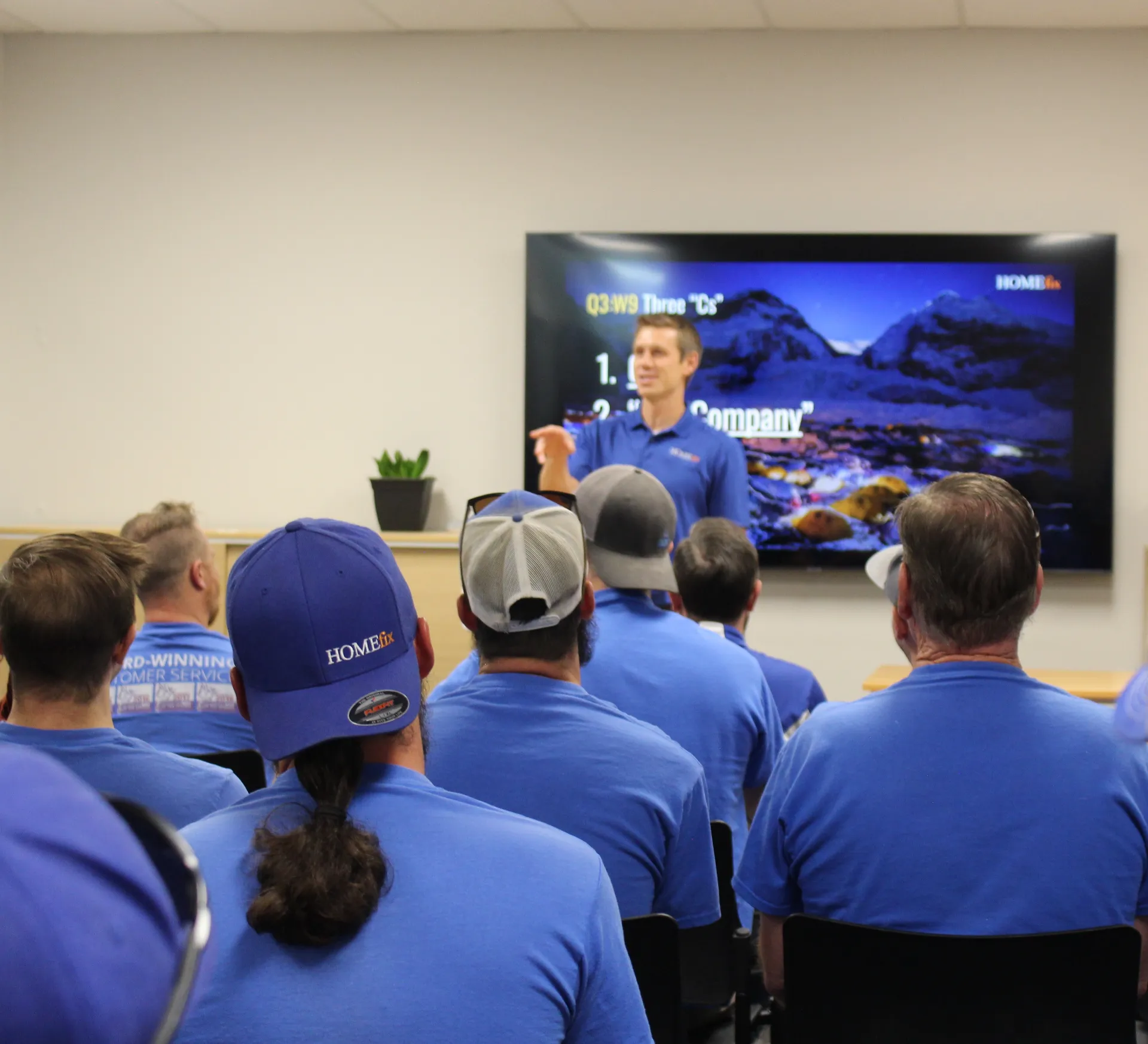 HOMEfix founder/owner in a blue shirt speaks to team wearing matching blue shirts and caps. A screen behind displays text titled "Three C's" with a mountain image.