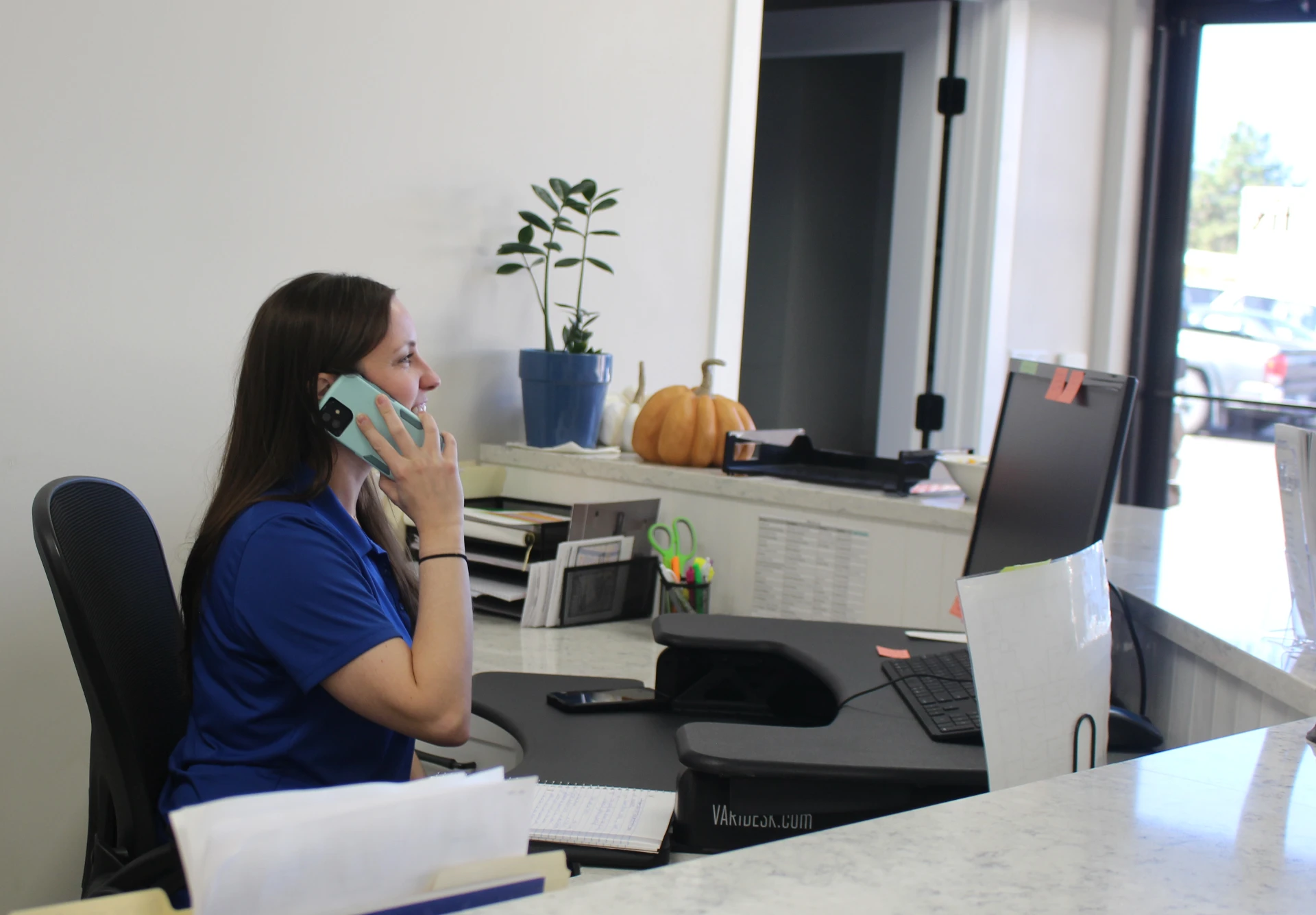 A woman in a blue shirt is at a reception desk, speaking on a phone. The desk has a computer, a plant, and office supplies. The atmosphere is professional.