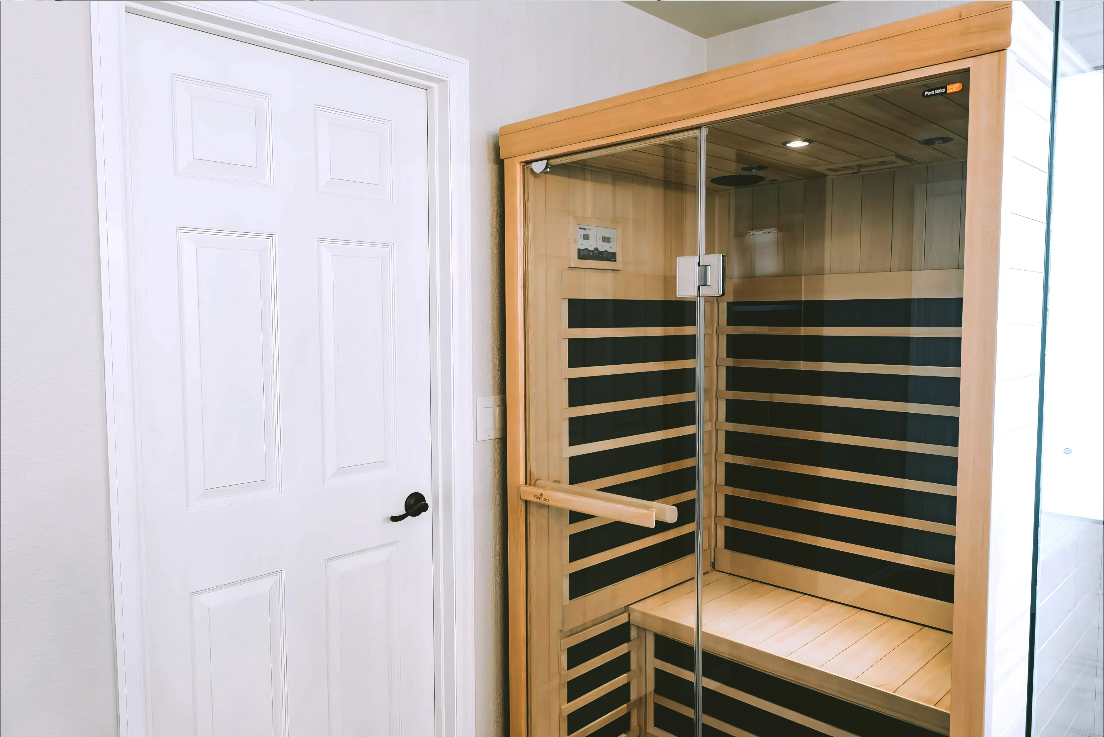 Modern wooden infrared sauna with glass door next to a white paneled door inside a room.