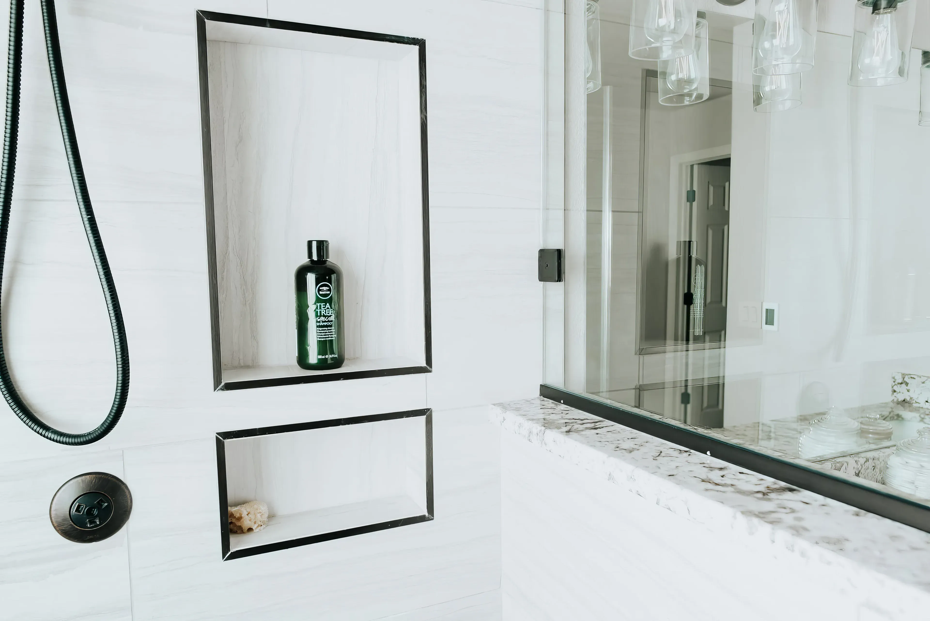 Close-up of a modern shower niche with a green bottle of tea tree shampoo and a sponge, next to a glass shower door and marble countertop.