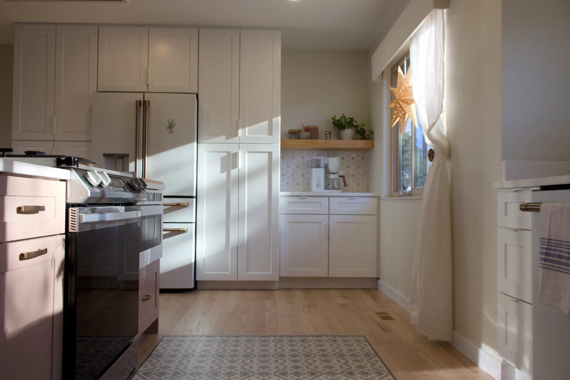 A serene kitchen with white cabinets, a modern oven, and wood flooring. Sunlight streams in through a window adorned with a star decoration.