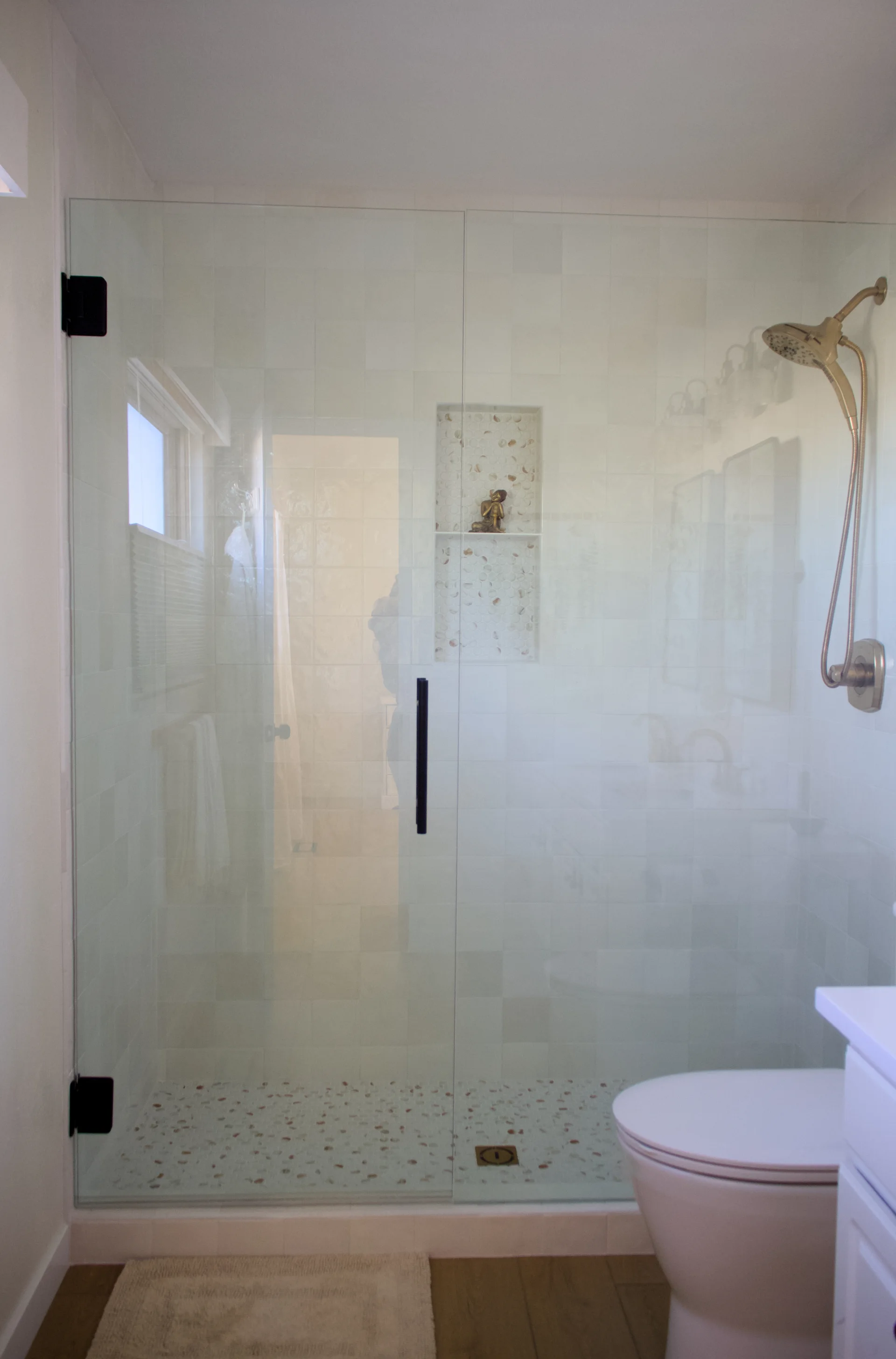Modern bathroom featuring a glass shower enclosure with a sleek black handle and gold fixtures. A small niche with decorative pieces is visible inside.