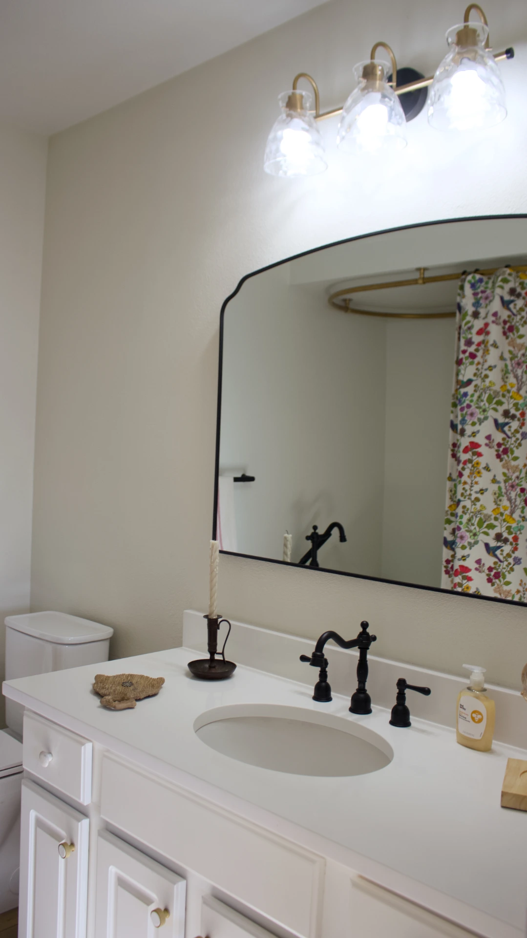 A bright bathroom with a white vanity, a black faucet, and a large mirror. Above are three modern lights. Colorful floral shower curtain in view.