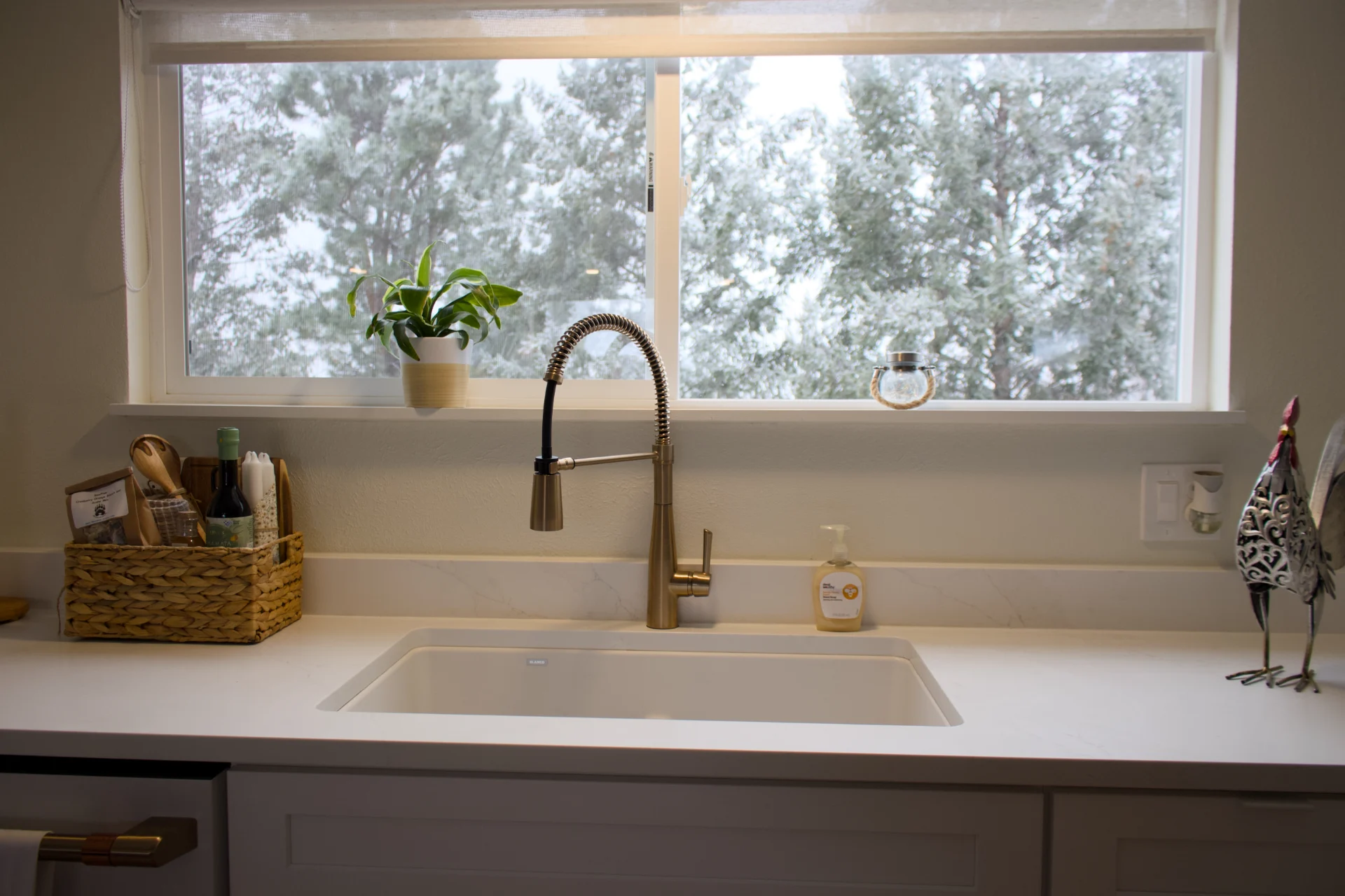 Bright kitchen with a gold faucet and white sink under a window showing snowy trees. A potted plant and basket sit on the counter, creating a cozy feel.