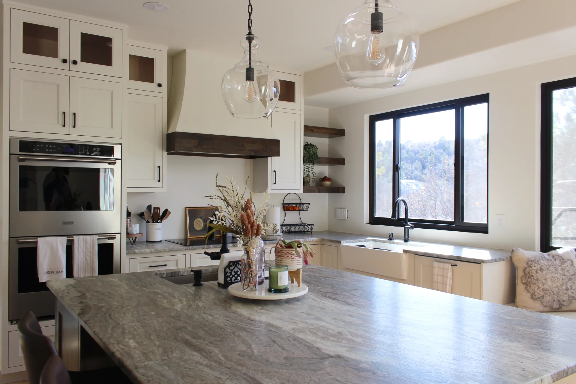 Bright kitchen with marble island, white cabinets, stainless steel double oven, farmhouse sink under large window, and decorative dried flowers on the island.
