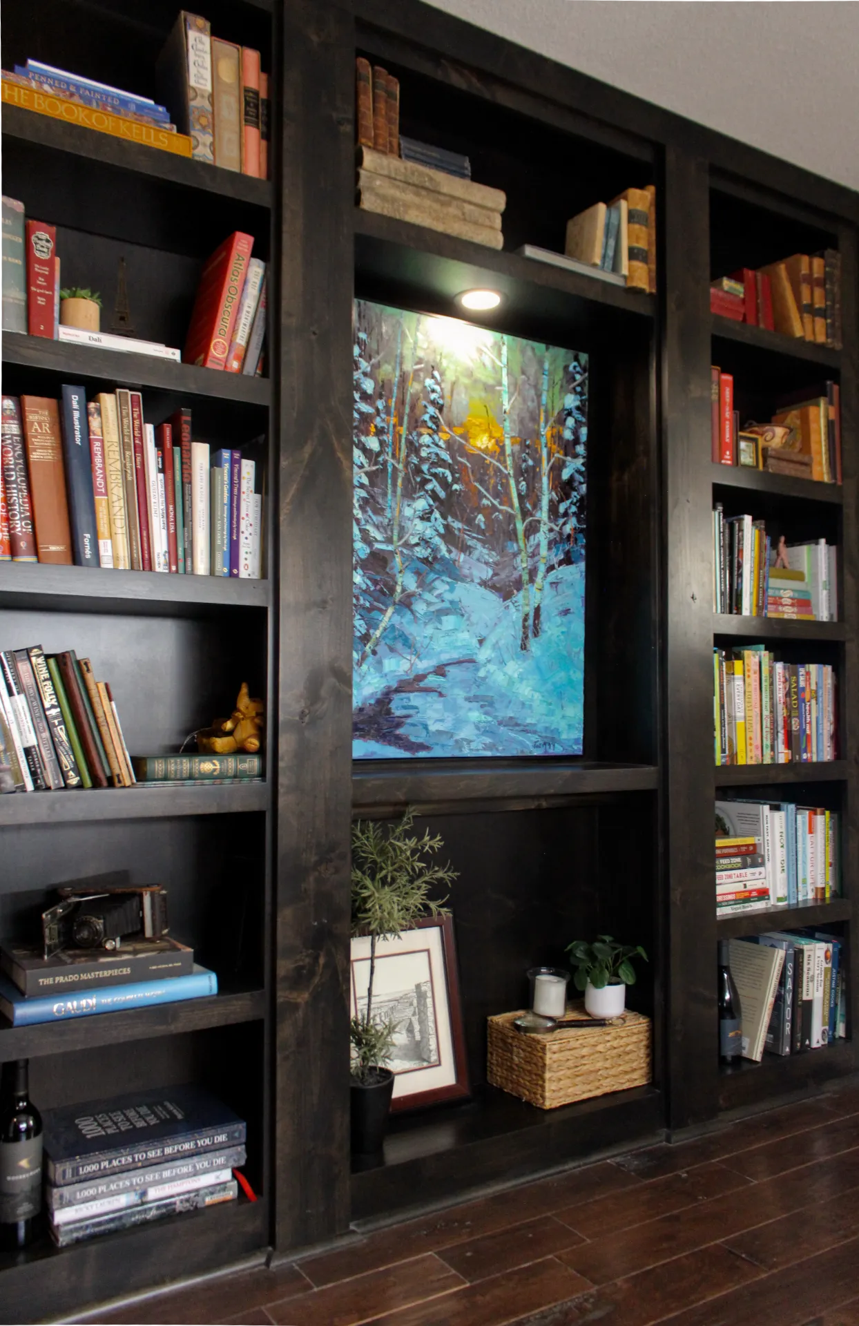 Dark wood built-in bookshelf filled with books, a framed picture, a basket, plants, and a painting of a snowy forest scene illuminated by a built-in light.