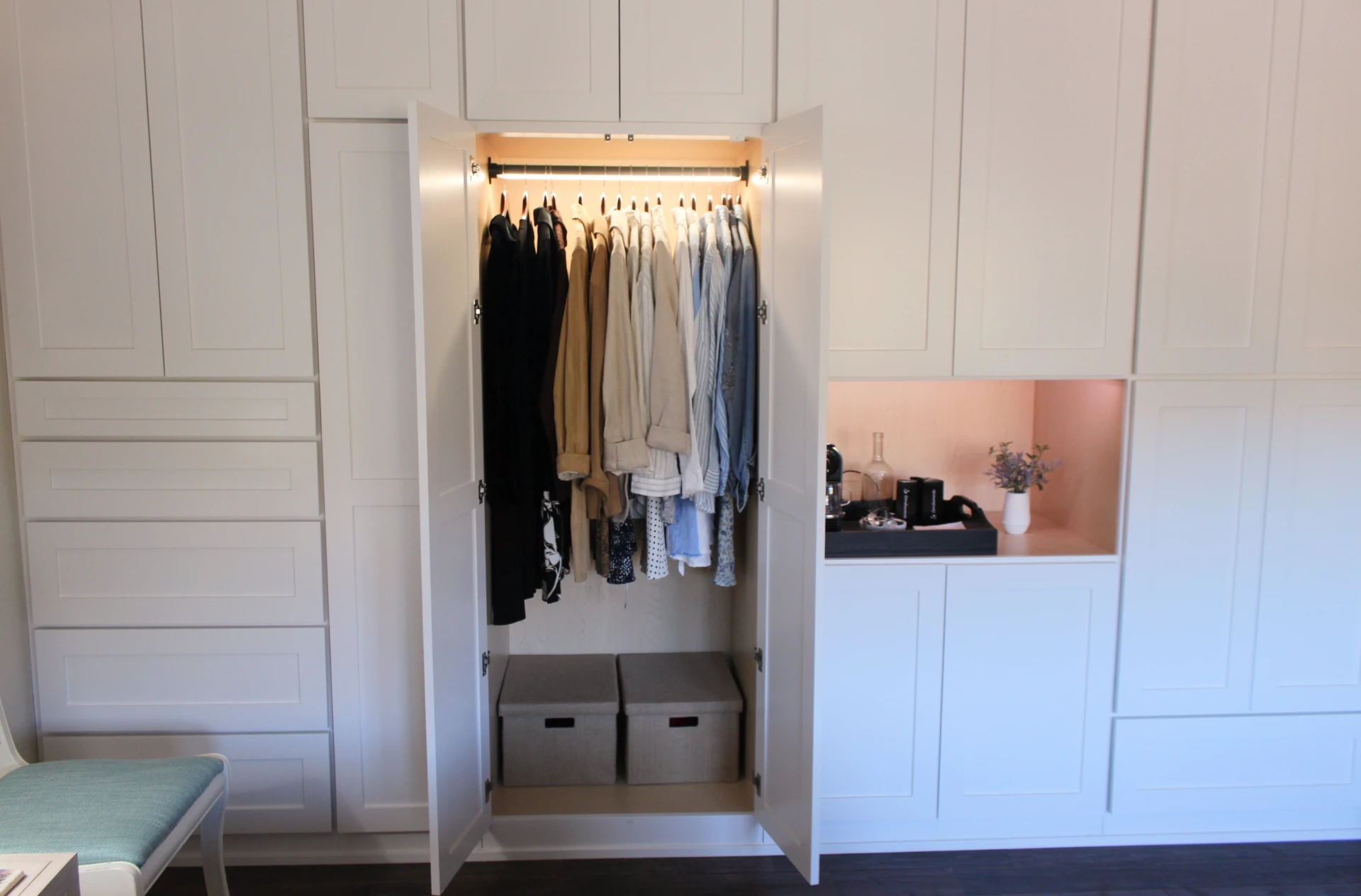 Open white wardrobe with hanging shirts in various shades and two fabric storage boxes below, next to white cabinets with a small built-in nook featuring a plant and coffee accessories.