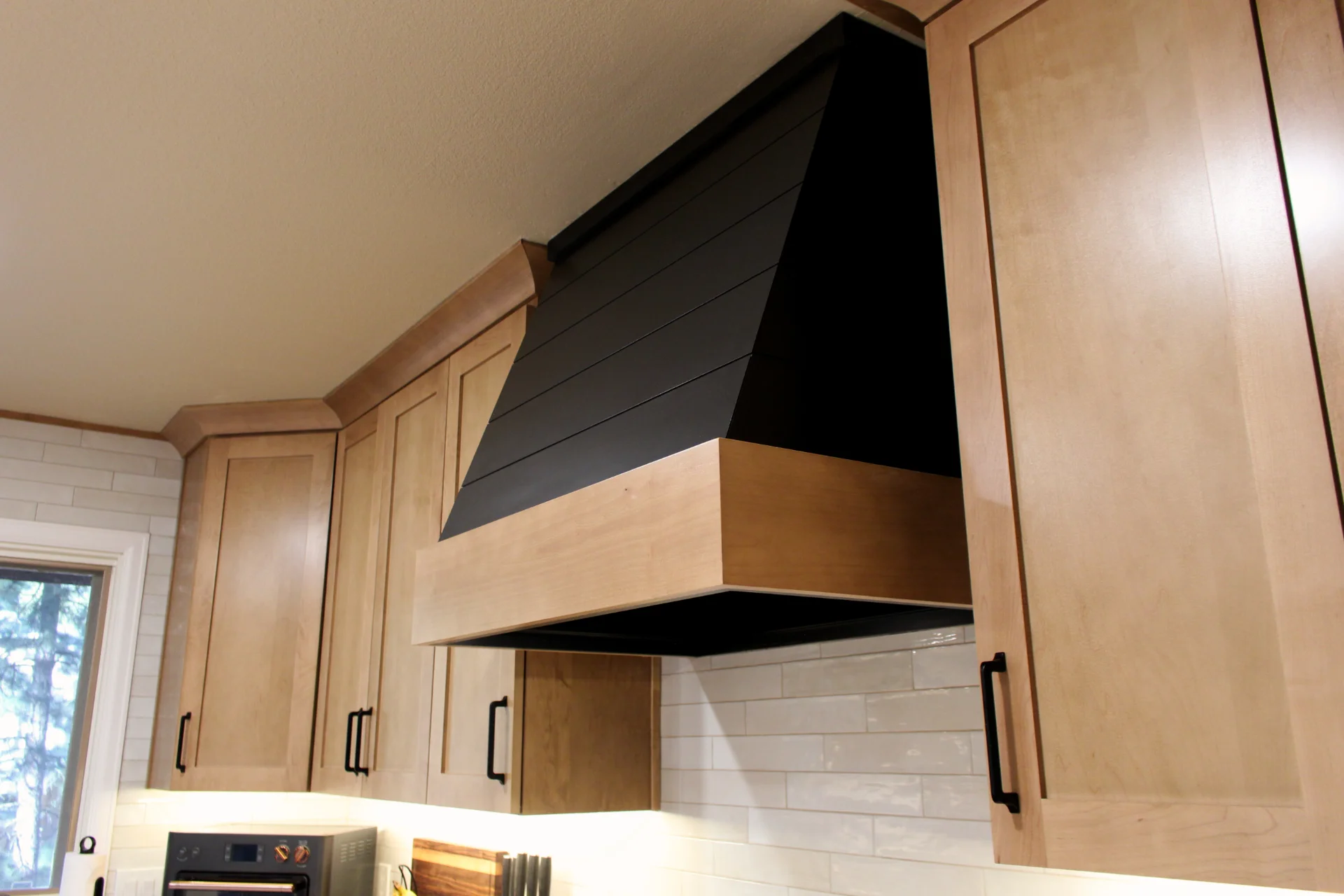 Black and wood custom kitchen range hood mounted above stove between light wood cabinets with black handles.