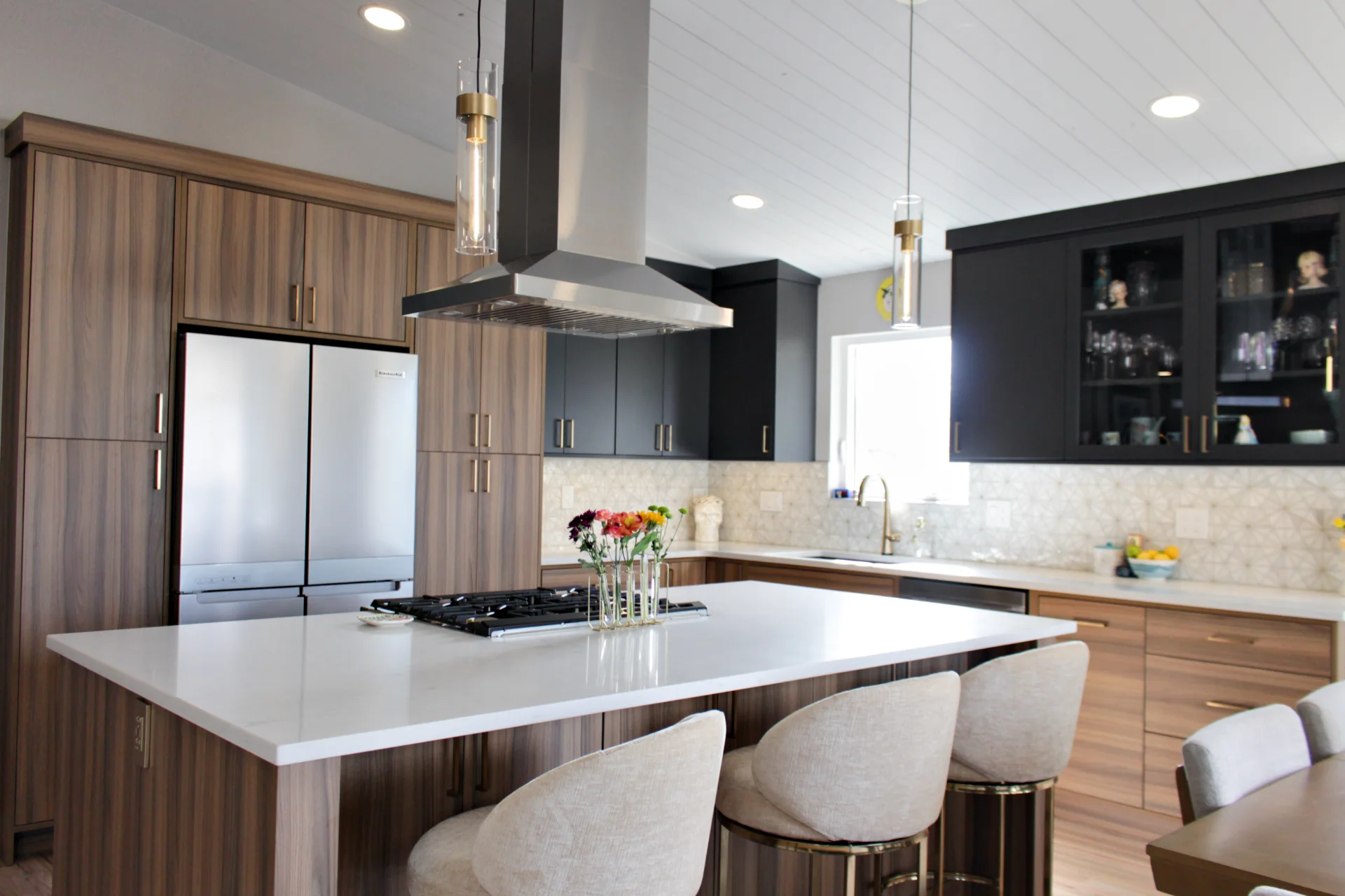 Modern kitchen with walnut and black cabinets, a large white island with beige stools, stainless steel refrigerator, and pendant lights.