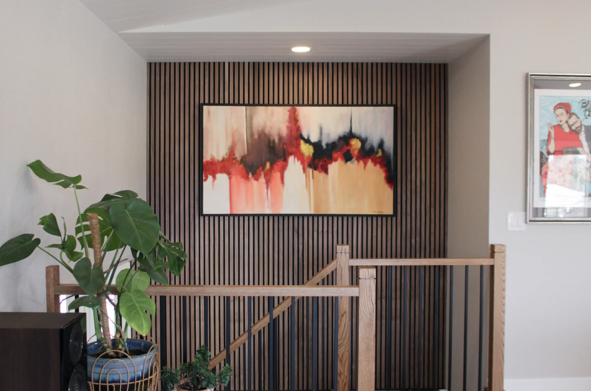 Modern interior stairwell with vertical wooden slat wall, abstract painting with red and beige tones, and large green potted plant.