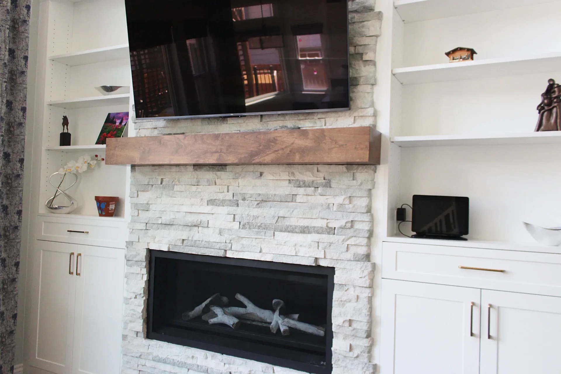 Modern living room with a white stone fireplace, wooden mantel, mounted flat-screen TV, and white built-in shelves on either side.