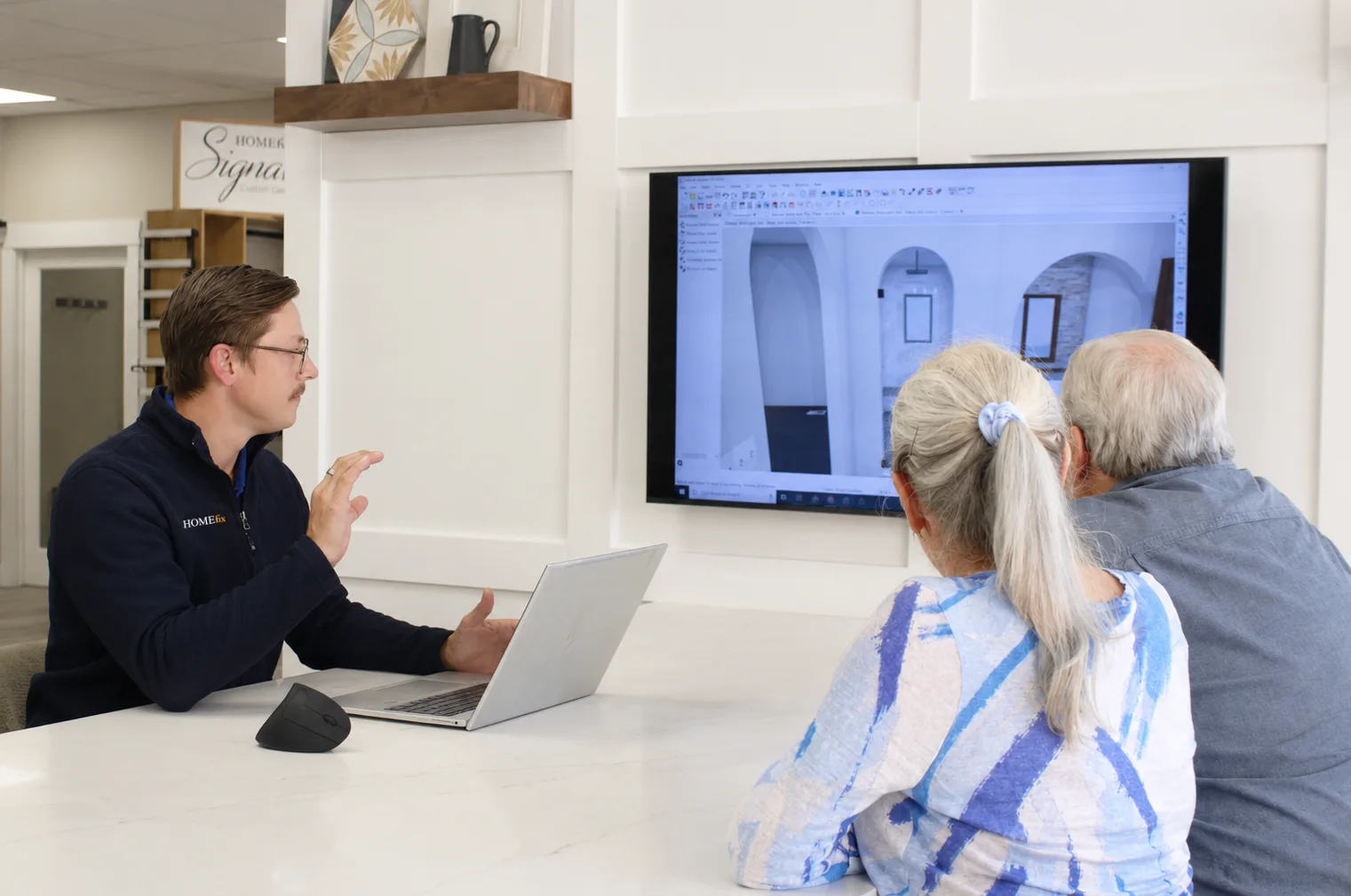 Man explaining home design on a laptop to an older couple watching a screen with architectural visuals.