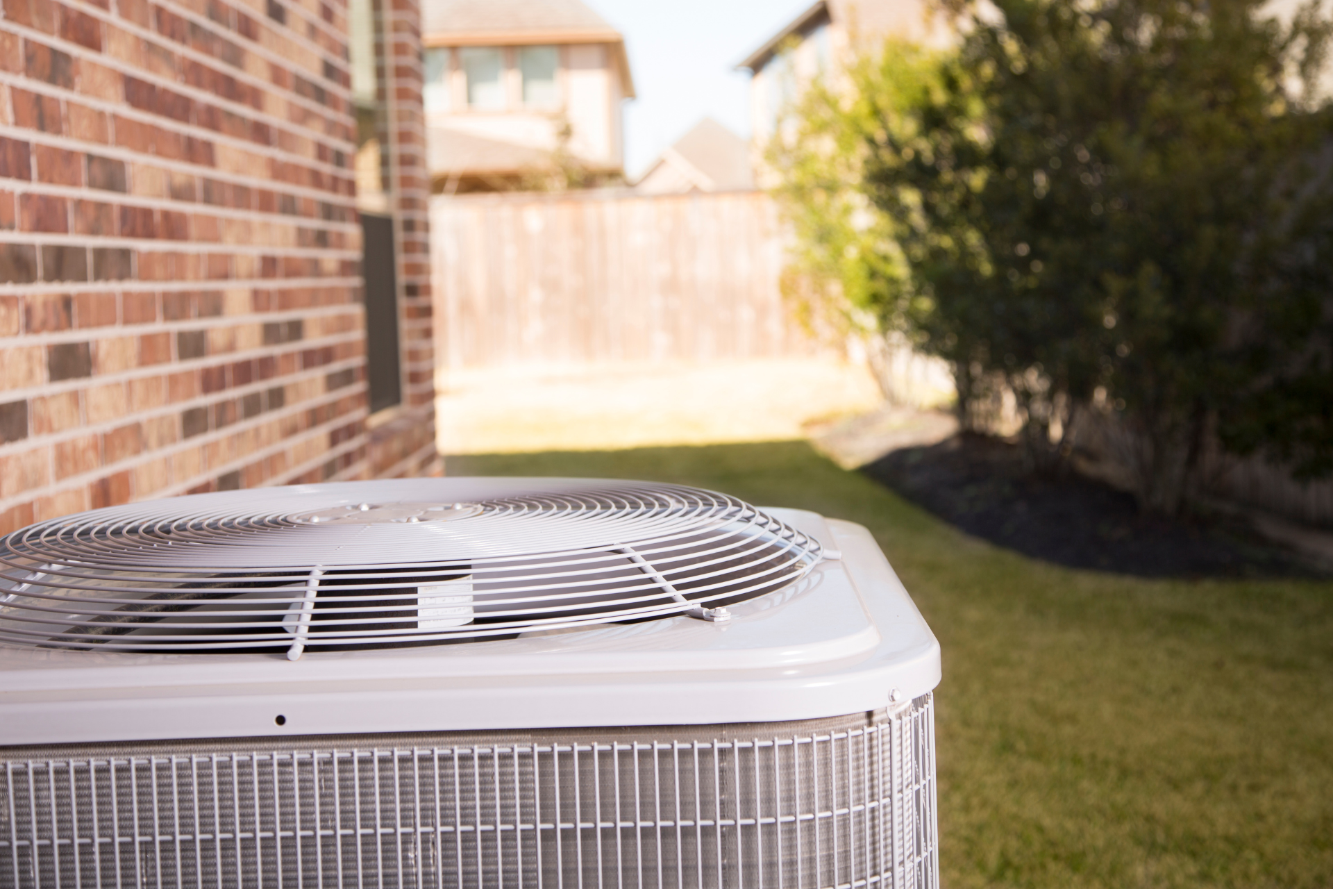 Outdoor air conditioning unit next to a brick house with a fenced backyard and green grass.