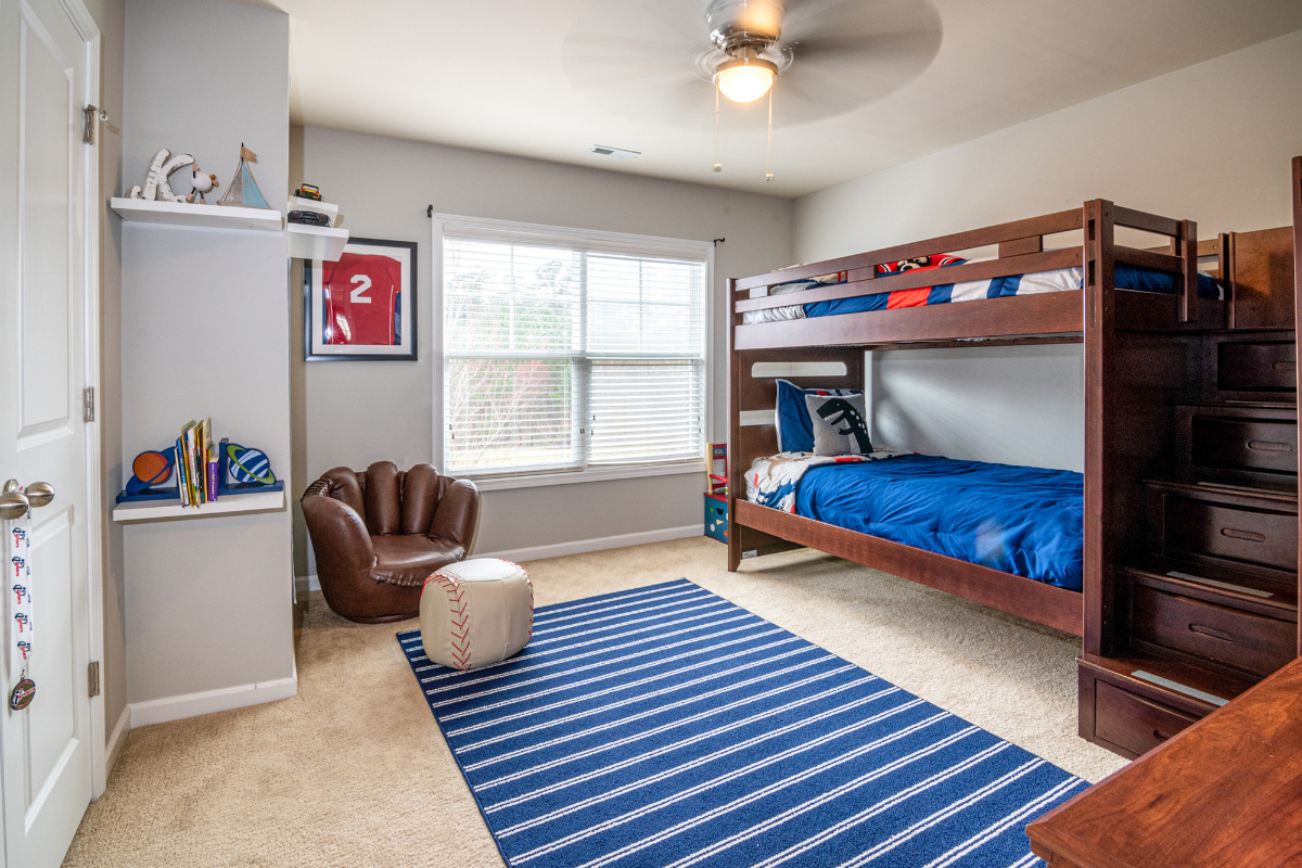 Kids' room with wooden bunk beds, blue beddings, striped rug, baseball chair, and window with blinds.