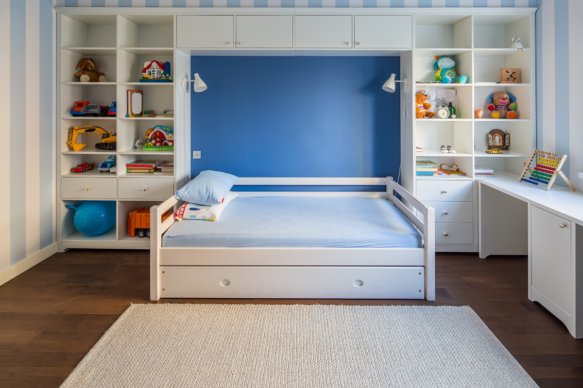 Child's room with white bed, blue pillow, built-in shelves holding toys, and a desk with an abacus.