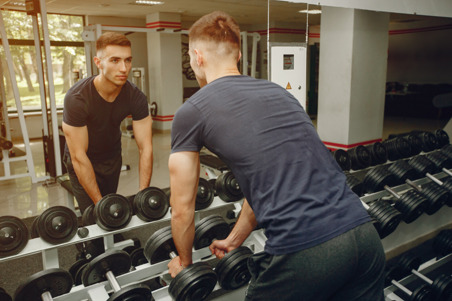 A man looking into the mirror while getting ready to lift a pair of dumbbells at the weight rack.