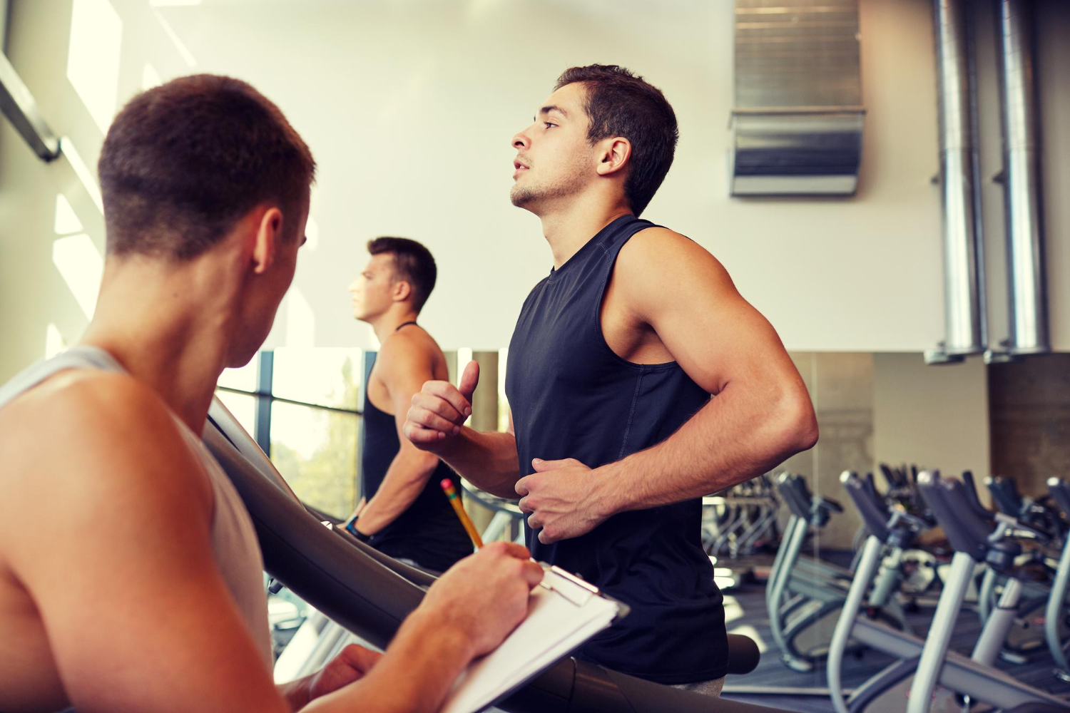 A man on a treadmill in a gym with another man writing down notes next to him.