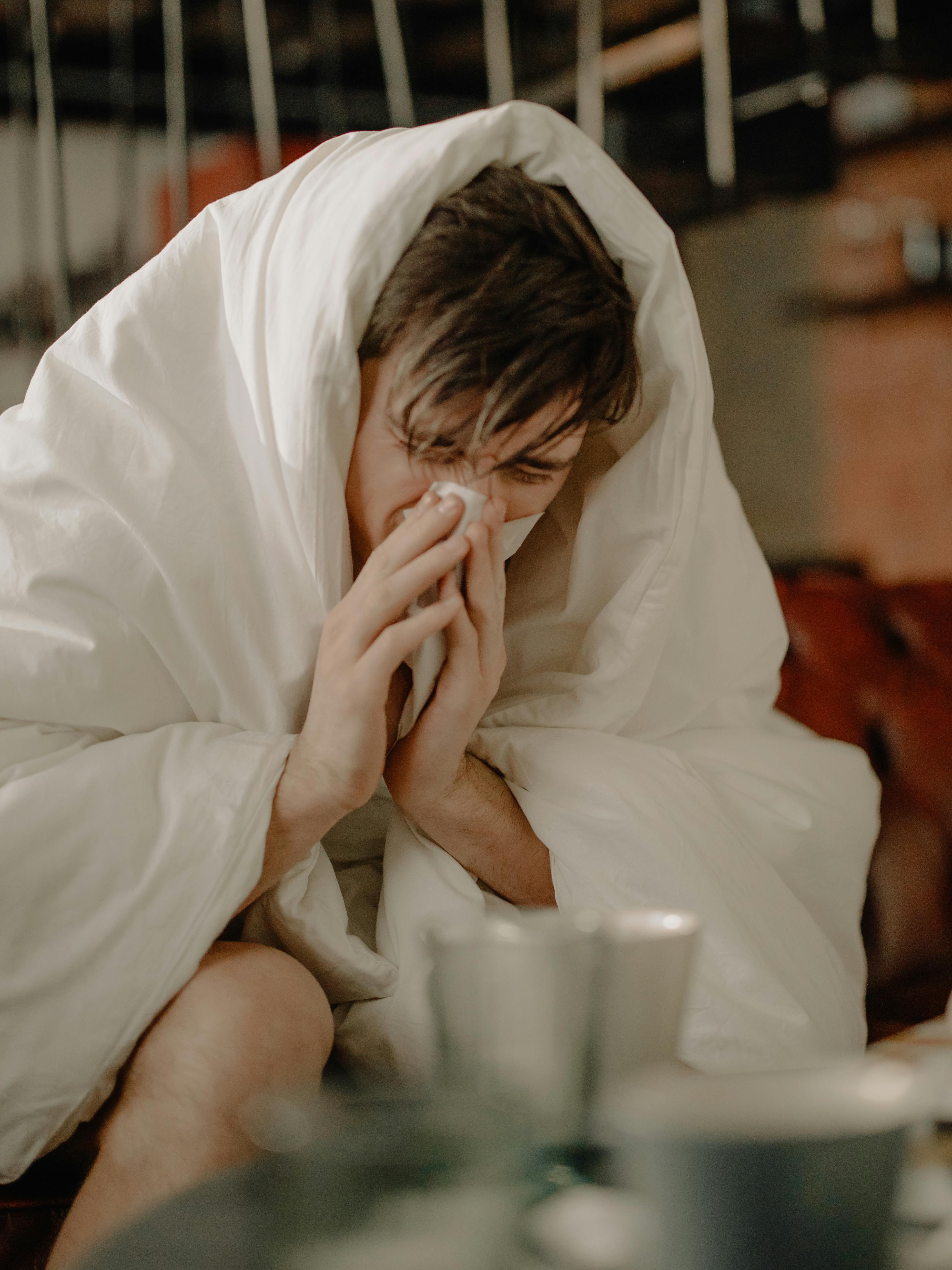 A man sneezing into a tissue with a blanket wrapped around him.
