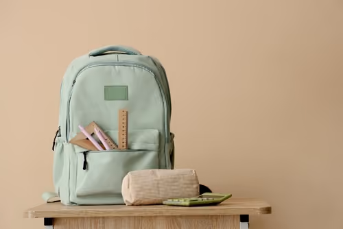 Light green backpack with wooden ruler and pink pencils in front pocket on a wooden table.