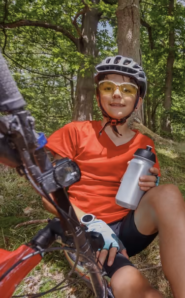 Boy in a helmet and sunglasses sitting on the ground with a bike and holding a water bottle in forest.