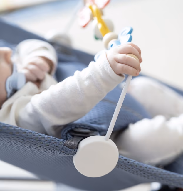 Baby lying on a blue fabric seat holding a rattle toy with a yellow handle and red wings.