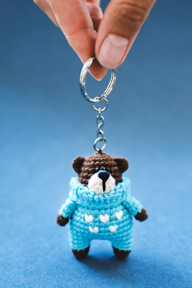 Hand holding a crocheted bear keychain wearing a blue sweater with white hearts.