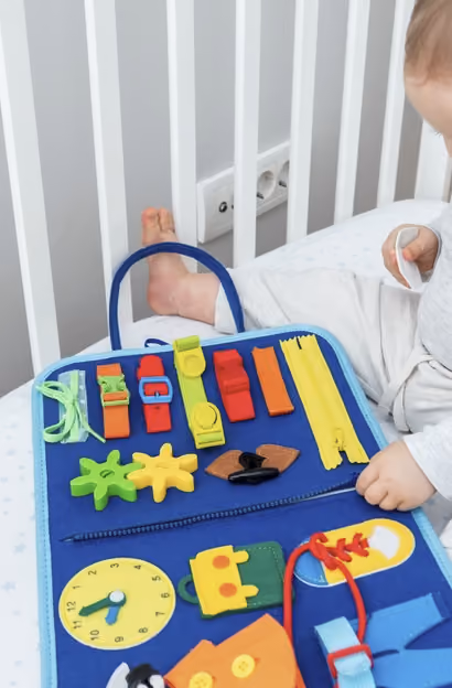 Baby playing with a blue activity board featuring buckles, zippers, laces, and a toy clock.