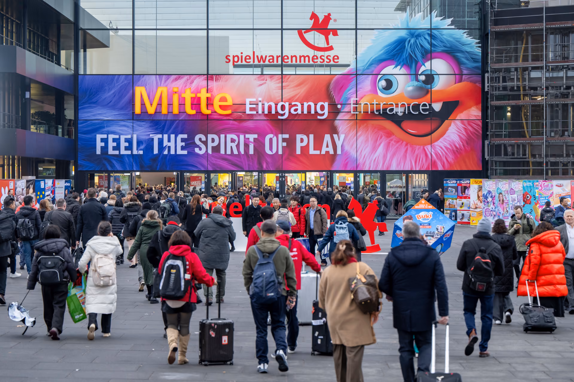 Crowd walking into Spielwarenmesse toy fair entrance with colorful furry character and signage.