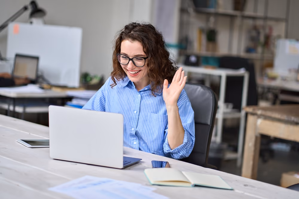Woman smiling and waving while video chatting on a laptop at a desk in an office setting.