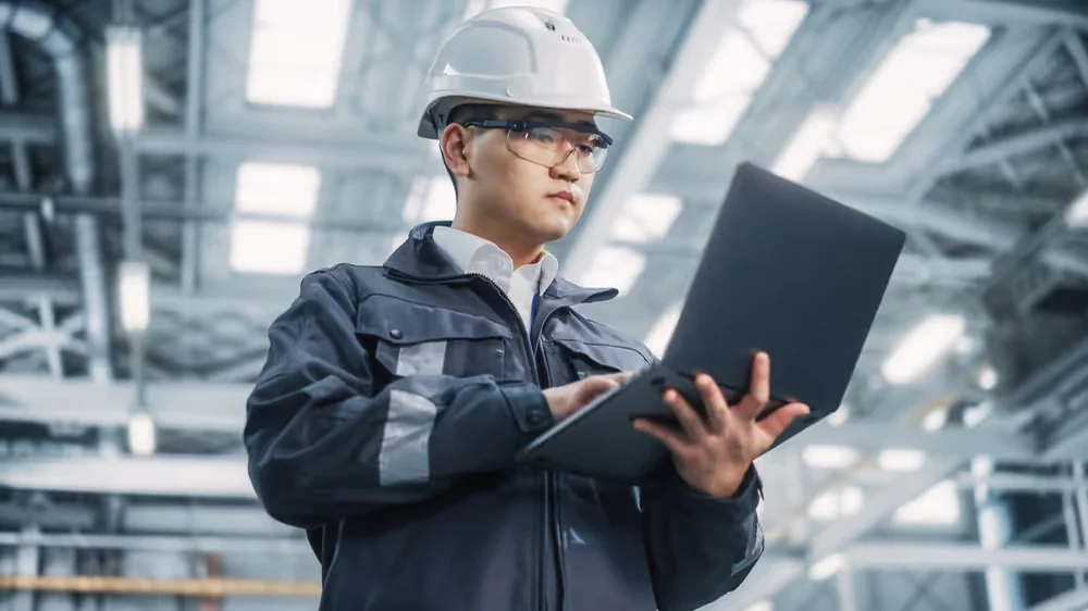 Worker using a portable computer beside production machinery.