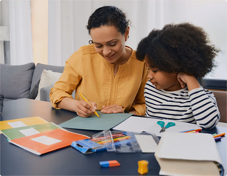 A woman and a child are sitting at a table with a book and a pile of crayons.