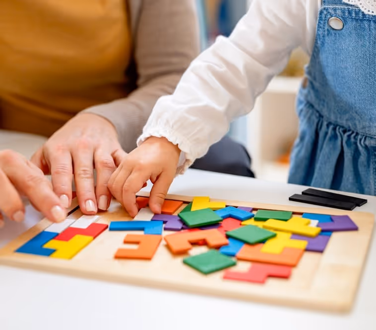 A child and an adult are seated at a table, playing with a colorful wooden tangram puzzle. The child's hand is reaching to place a green piece on the board, while the adult's hand is nearby, assisting.