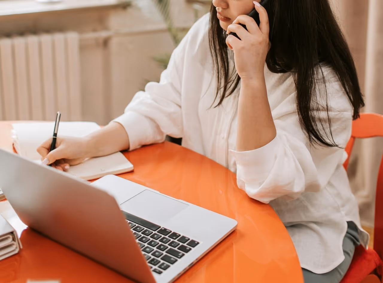 A person wearing a white shirt is sitting at a bright orange table, using a laptop, and writing in a notebook while holding a phone to their ear.