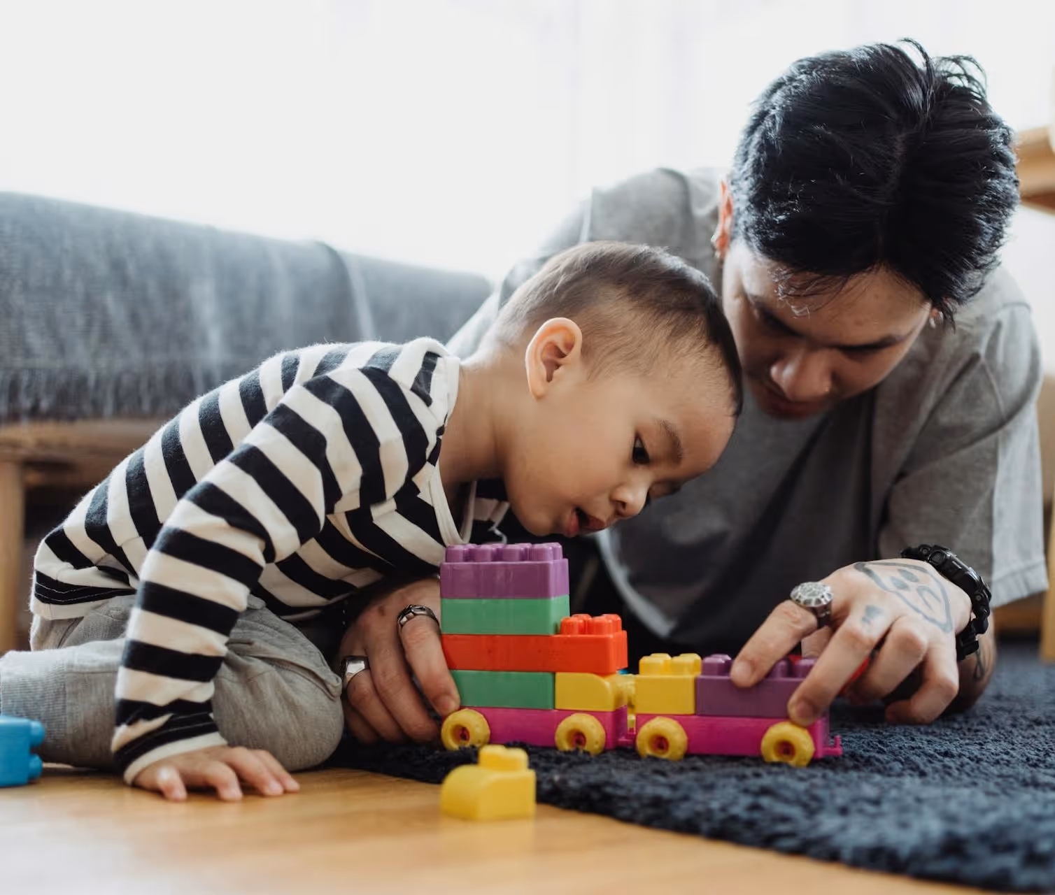 A bcba and a young child are lying on a rug, building a colorful toy train with plastic building blocks. The bcba is looking at the child, who is focused on the toy.