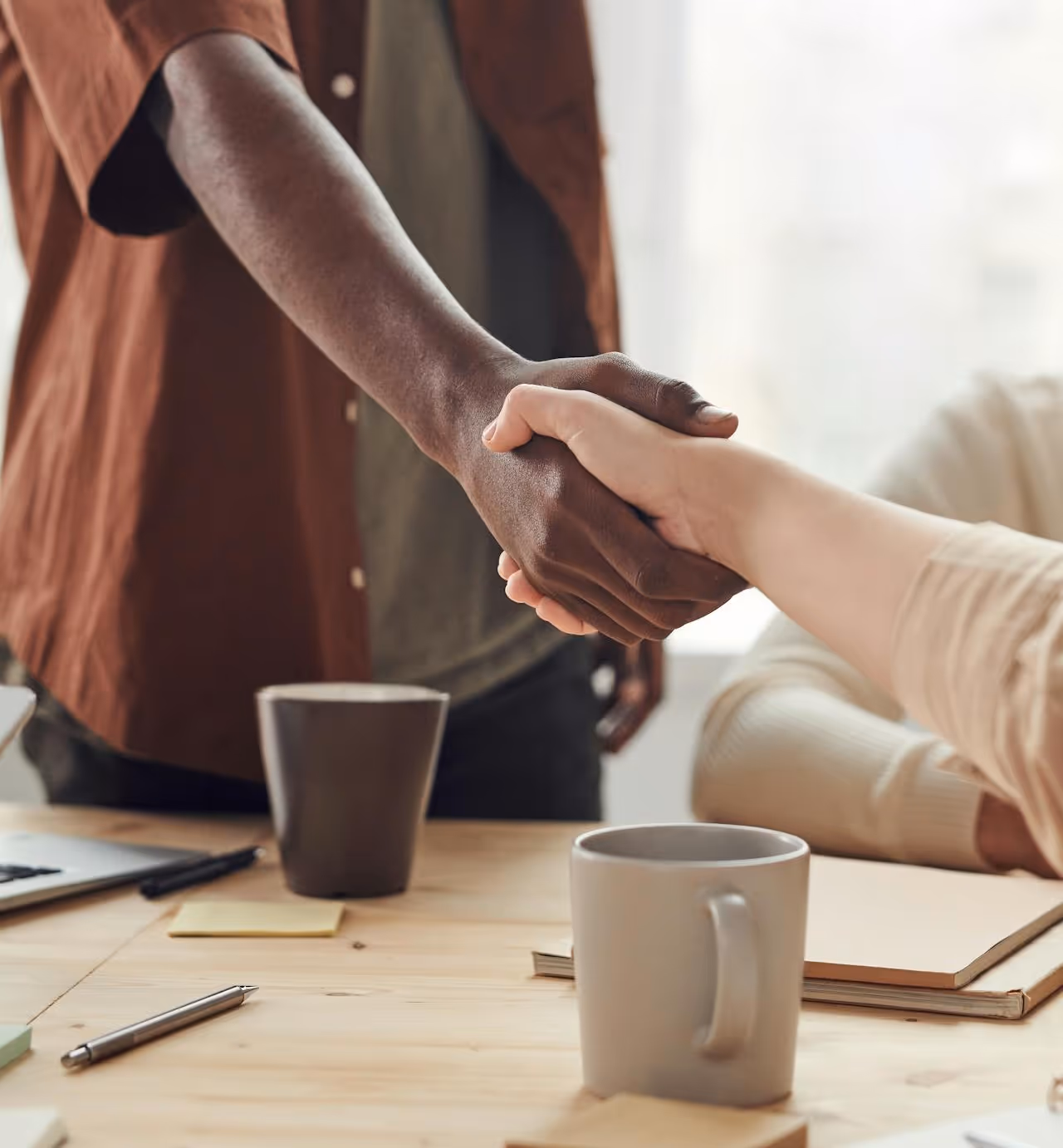 Two people are shaking hands over a wooden desk, with coffee mugs and other office supplies visible. One person has dark skin and the other has light skin.