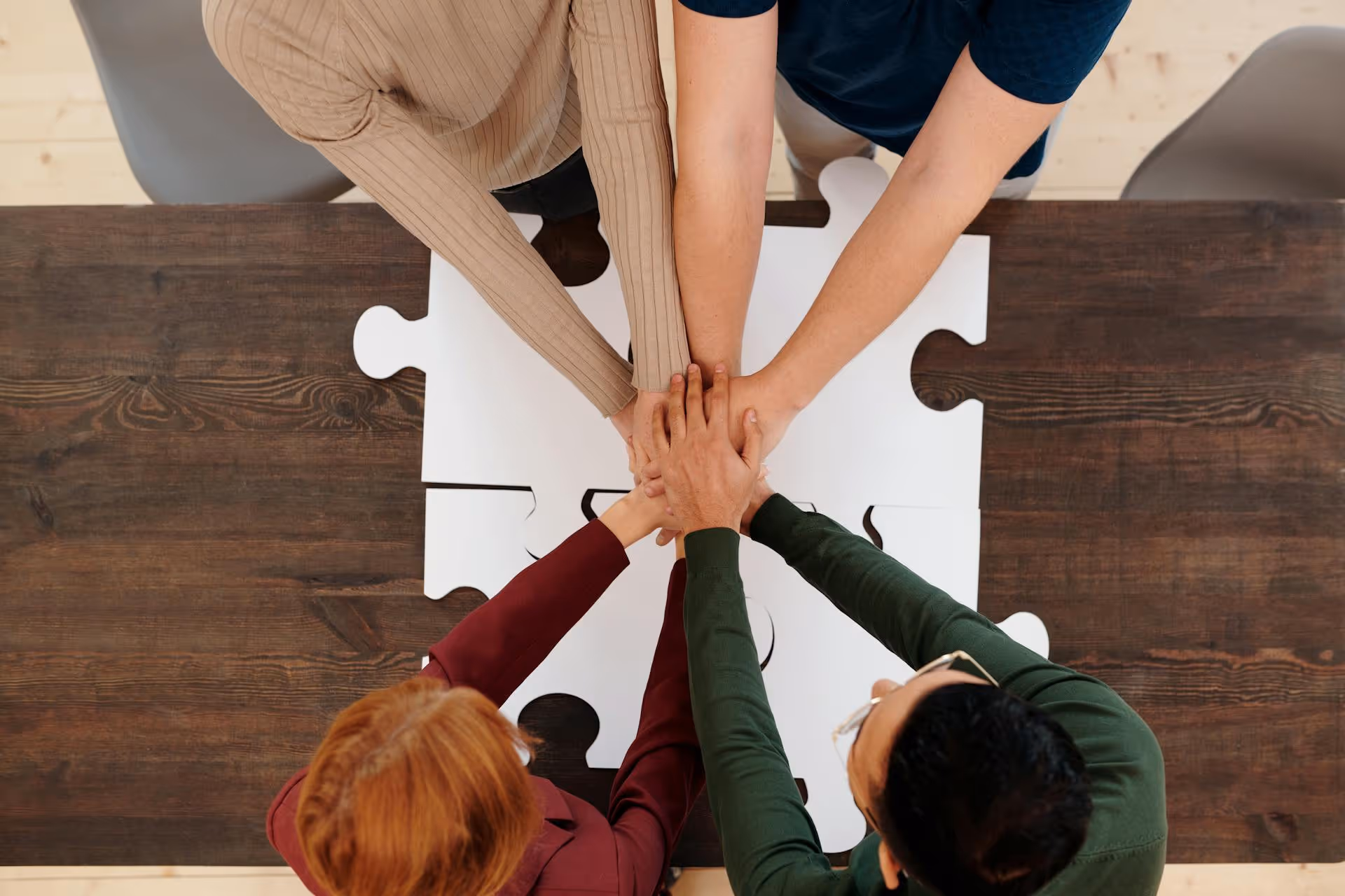 A diverse group of four people with their hands stacked in the center, symbolizing teamwork and unity. They are gathered around a table with a large, white jigsaw puzzle placed on it.