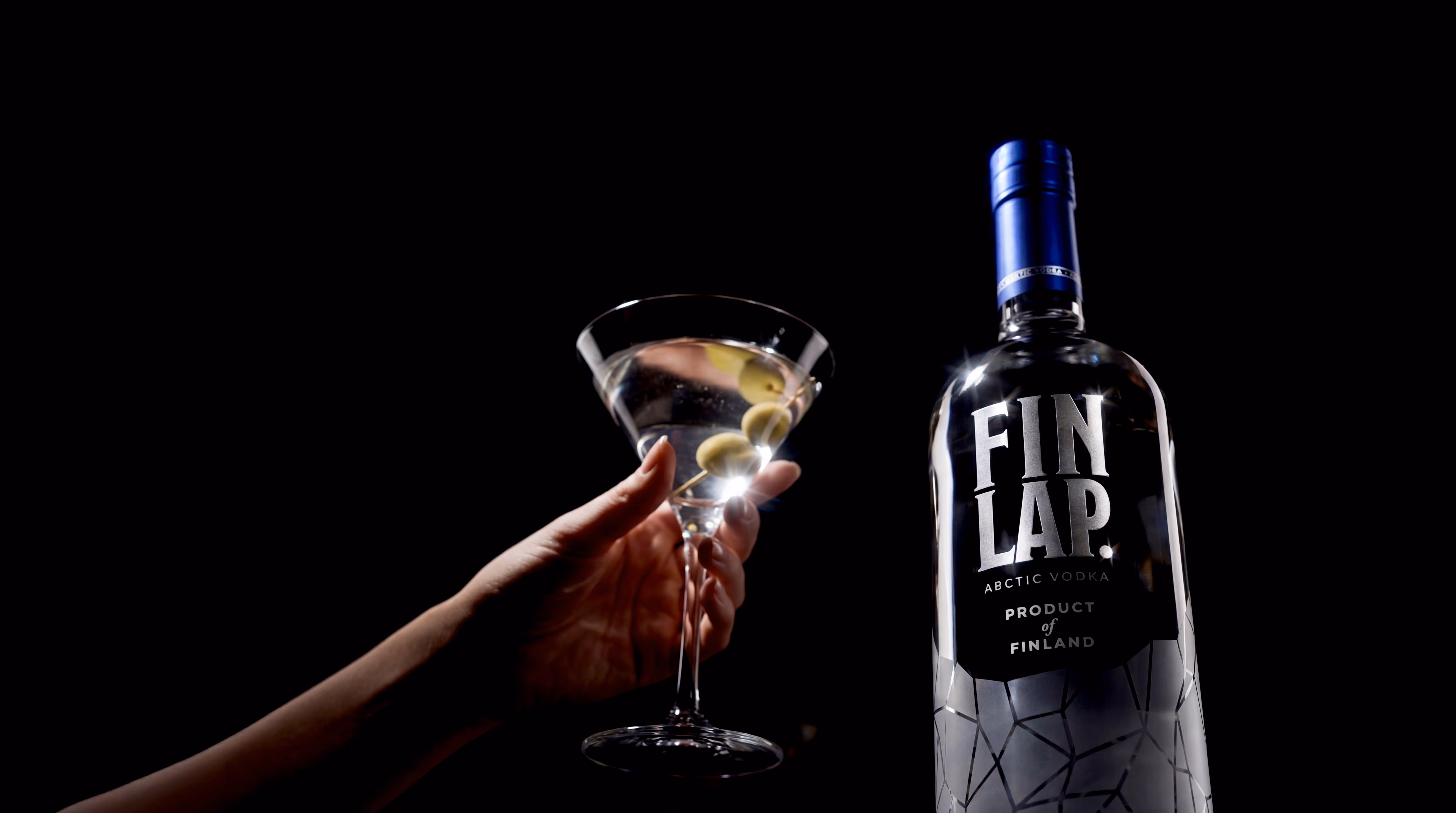 Hand holding a martini glass with olives next to a bottle of FIN LAP Arctic Vodka against a black background.