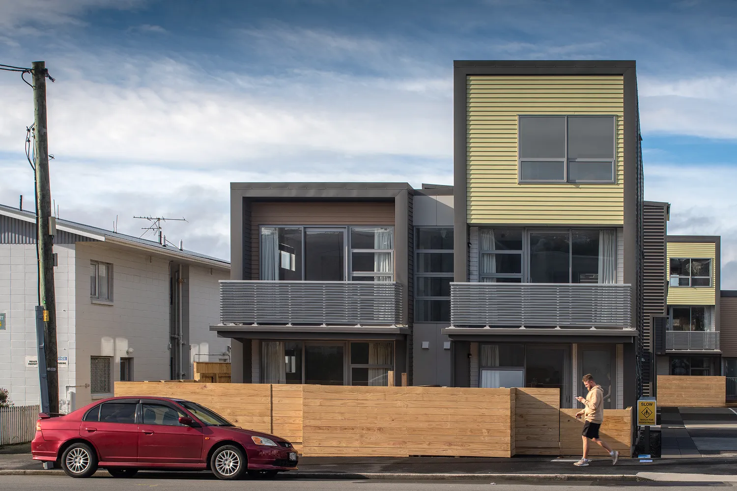 Two storey townhouses with dark grey and yellow cladding and horizontal slat balustrades on upper level balconies, with timber fencing and a red car parked on the street