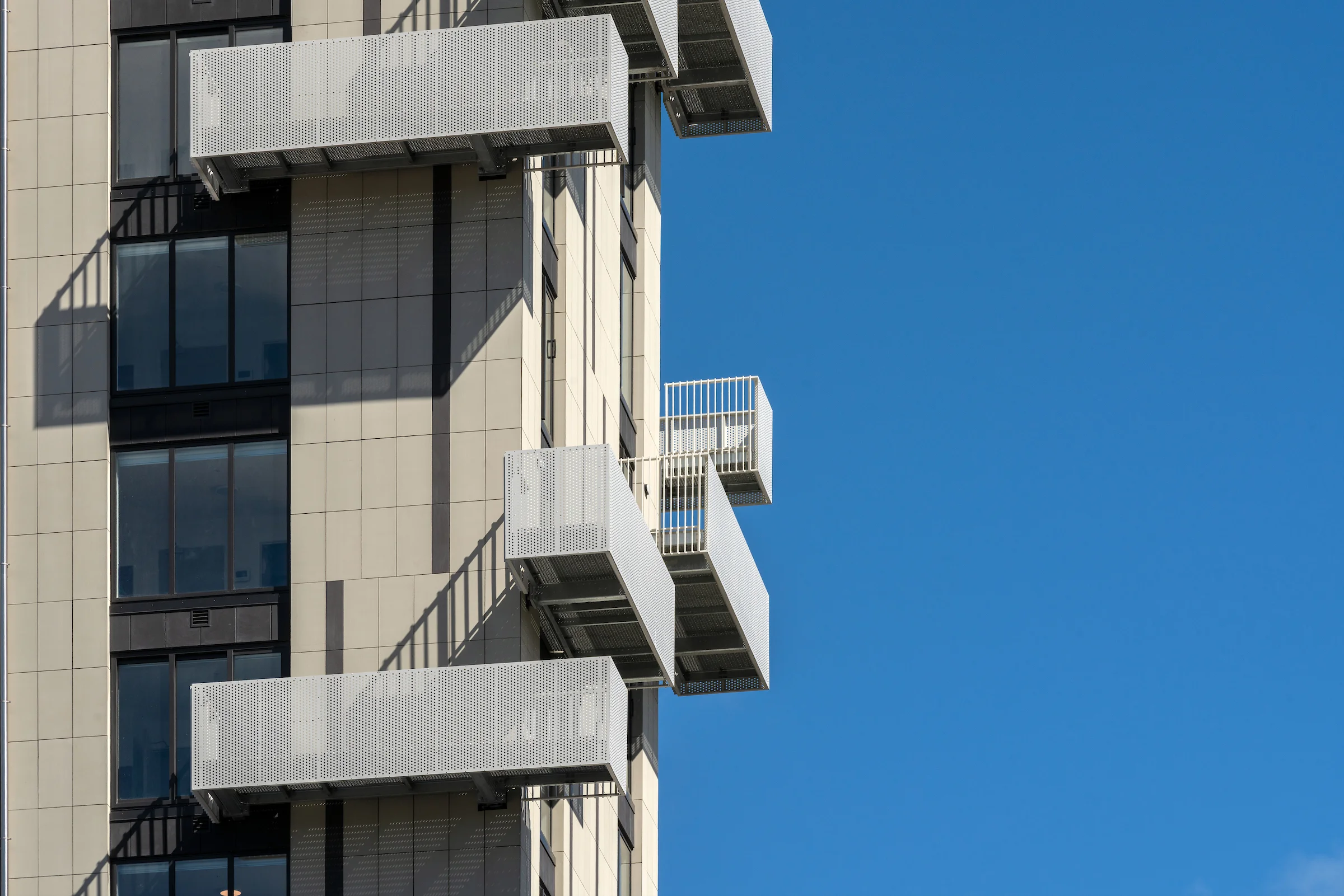 Close up of white perforated metal balconies and slim white balustrades projecting from a high rise building facade against a clear blue sky