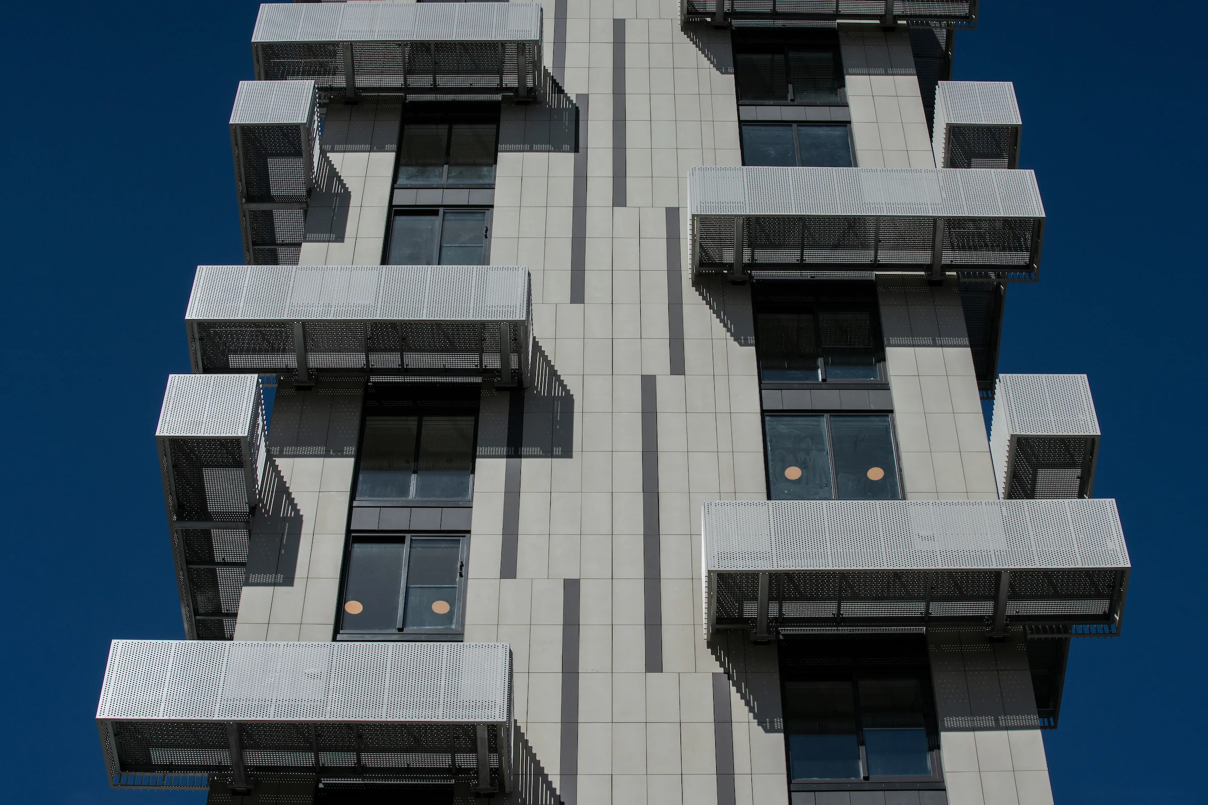 Upward view of staggered perforated metal balconies with slim black balustrades on a high rise building facade